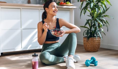 Shot of athletic woman eating a healthy bowl of muesli with fruit sitting on floor in the kitchen at home